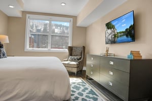Black chest of drawers, flat screen TV and upholstered sitting chair next to the large window with mountain views.