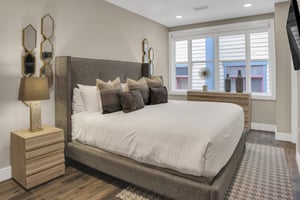 A grey upholstered kingsize bed with grey throw pillows, mirror art on the wall, large window, and serenecolored walls.