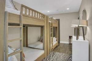 Two light colored wood bunkbeds, pushed end to end with a grey and white floor rug and wood floors.
