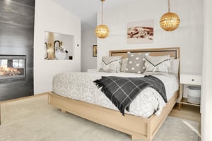 The light colored wood kingsized bed has grey and white pattened bedspread and throw pillows, and a dark grew throw blanket.