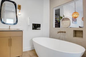 Elegant standalone soaking tub in the main bathroom with gold wall faucet, wood vanity, and black framed mirror.