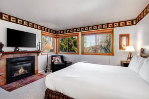 Snow Flowers main suite has a king size bed with white linens, fireplace and TV, blue chair under 3 windows and floor lamp.