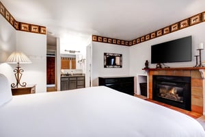 Snow Flowers main suite has a king size bed with white linens, attached bathroom, black dresser, and fireplace and TV above.