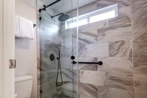 The walkin shower has grey and white patterned wall tile, black hardware, white toilet, and a white folded hanging towel.