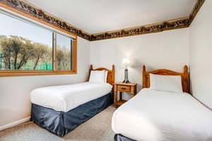 Two twin beds with medium toned wood headboards, blue bed skirt, and a nightstand and light in between them.