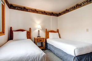 Two twin beds with medium toned wood headboards, blue bed skirt, and a nightstand and light in between them.