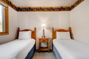 Two twin beds with medium toned wood headboards, blue bed skirt, and a nightstand and light in between them.