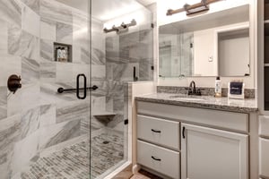 The shared bathroom has grey cabinets, 1 sink vanity, large mirror and light, and a walkin shower with grey and white tile.