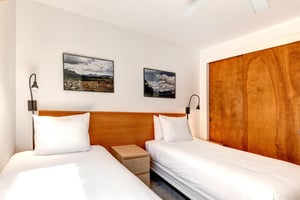 2 twin beds with crisp white linens, wood headboard with 2 attached lights, shared night stand and two pictures on the wall.