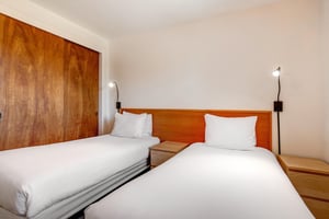 2 twin beds with crisp white linens, wood headboard with 2 attached lights, shared night stand and two pictures on the wall.