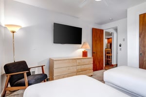2 twin beds with crisp white linens, wood dresser, wall mounted TV, and a blue chair with wooden arms.