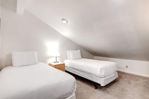 2 twin beds with crisp white linens, shared night stand with a lamp, and a pitched ceiling with recessed lighting.