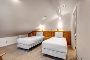 2 twin beds with crisp white linens, shared night stand, 2 lights, a pitched ceiling with recessed lighting and wooden pony wall.