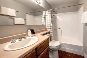 A bathroom with wooden cabinets, cream colored counter, single sink, white toilet and bathtubshower combination.