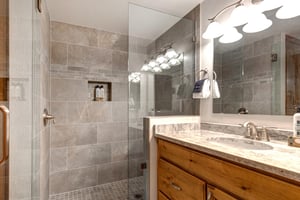 Bathroom with grey tiled shower and glass partition, one sink vanity with warm wood cabinets, large mirror, and light.