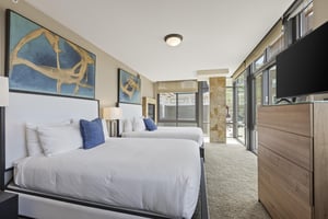 The second bedroom has two queen beds, crisp white linens, with blue throw pillows and modern art above the bed.