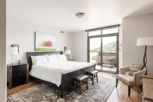 Main suite king bed, white linens and duvet, wooden headboard, hardwood floors with large Persian area rug, and sliding patio doors to access private deck