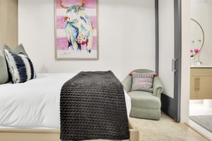 Guest bedroom with a colorful bull painting, a bed with crisp white linens and grey throw blanket, and a sitting chair.