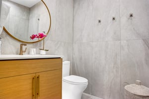 Ensuite bathroom with granite wall tiling, wooden cabinetry, round mirror, white counters, and white toilet.