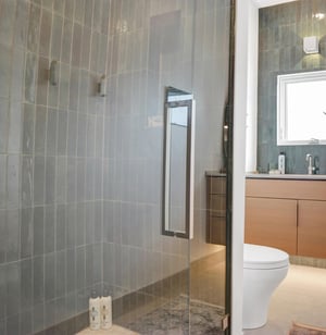 The bathroom has a walk in shower with deep green tile surround and glass doors and wood vanity with deep green tile backsplash.