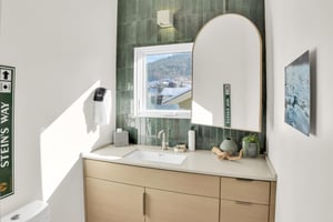 The bathroom has a wood vanity with deep green tile backsplash, window overlooking the mountain and town, and large mirror.