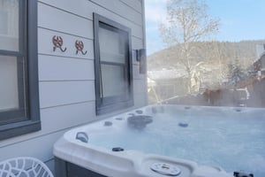 A steamy hot tub on the back deck against the house with a snowy mountain and town backdrop.