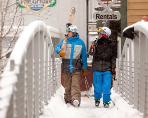 Skiers navigate a snowy bridge in the winter ski area, with equipment rental services visible nearby.