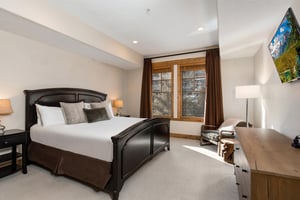 Relax in this comfortable bedroom featuring a dark wood bed with crisp linens, natural light from large windows, and modern amenities including a TV and workspace area.