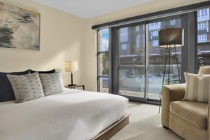 A king bed with crisp white linens, end tables with lamps, grey throw pillows, tan chair, floor lamp, and large windows.