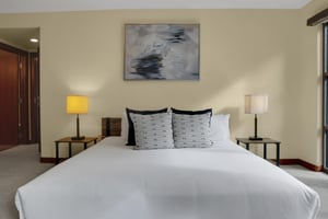 A king bed with crisp white linens, end tables with lamps, grey throw pillows, and abstract art above the bed.