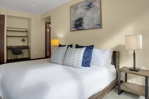 A king bed with crisp white linens, end tables with lamps, grey throw pillows, and small desk, task light and black chair.