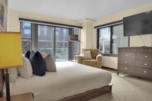 A king bed with crisp white linens, grey throw pillows, tan chair, floor lamp, wood dresser and flat screen TV.