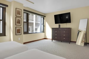 The end of two twin beds with a wooden dresser and wall mounted TV, large picture window and cream colored carpet.