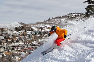 A skier dressed in orange and red at Park City Mountain Resort on a steep snowy slope with the town behind him.