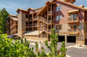 A welcoming mountain retreat with warm wood and stone architecture, featuring multiple balconies, convenient covered parking, and beautifully landscaped surroundings under a clear blue sky.