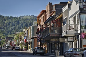 A charming mountain town main street lined with historic buildings, local shops, and eateries, set against a backdrop of lush forested hillsides and clear blue skies.