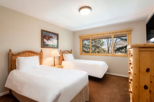 Bright guest bedroom with two beds, wood headboards, bedside lamp and large windows overlooking trees