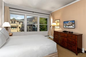 Bright bedroom with queen bed, wood dresser, TV and large windows overlooking the surrounding mountain area.