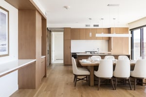 Dining room with linear chandelier light, kitchen in the background and wooden cabinetry.