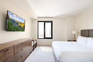 King bed in crisp white linens, picture window, wooden dresser, and wall mounted TV.