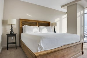 King bed with white linens, wood headboard and bedside lamp in a bright hotelstyle bedroom.