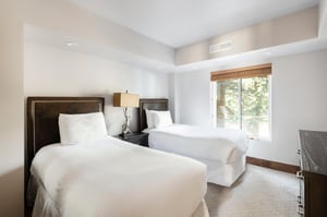 Bright bedroom with two beds, white linens, bedside table and window overlooking the trees.