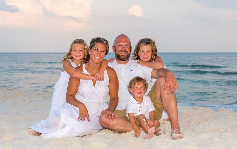 Smiling family of five sitting together on the sand at the beach with the ocean behind them during a sunset portrait session, wearing light coastal outfits.