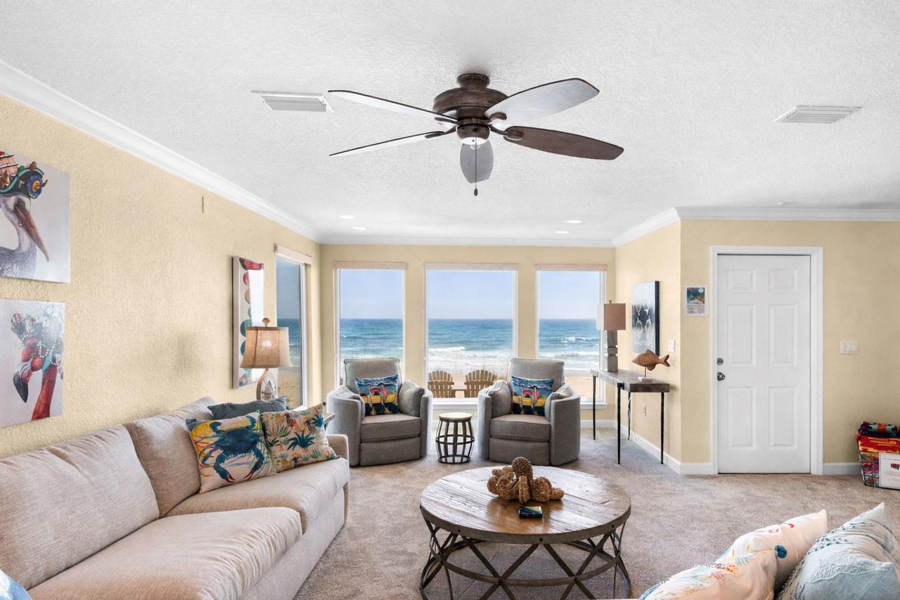 Your beachfront living room captures endless Gulf views through floortoceiling windows, where gentle ceiling fan breezes complement the coastal décor.