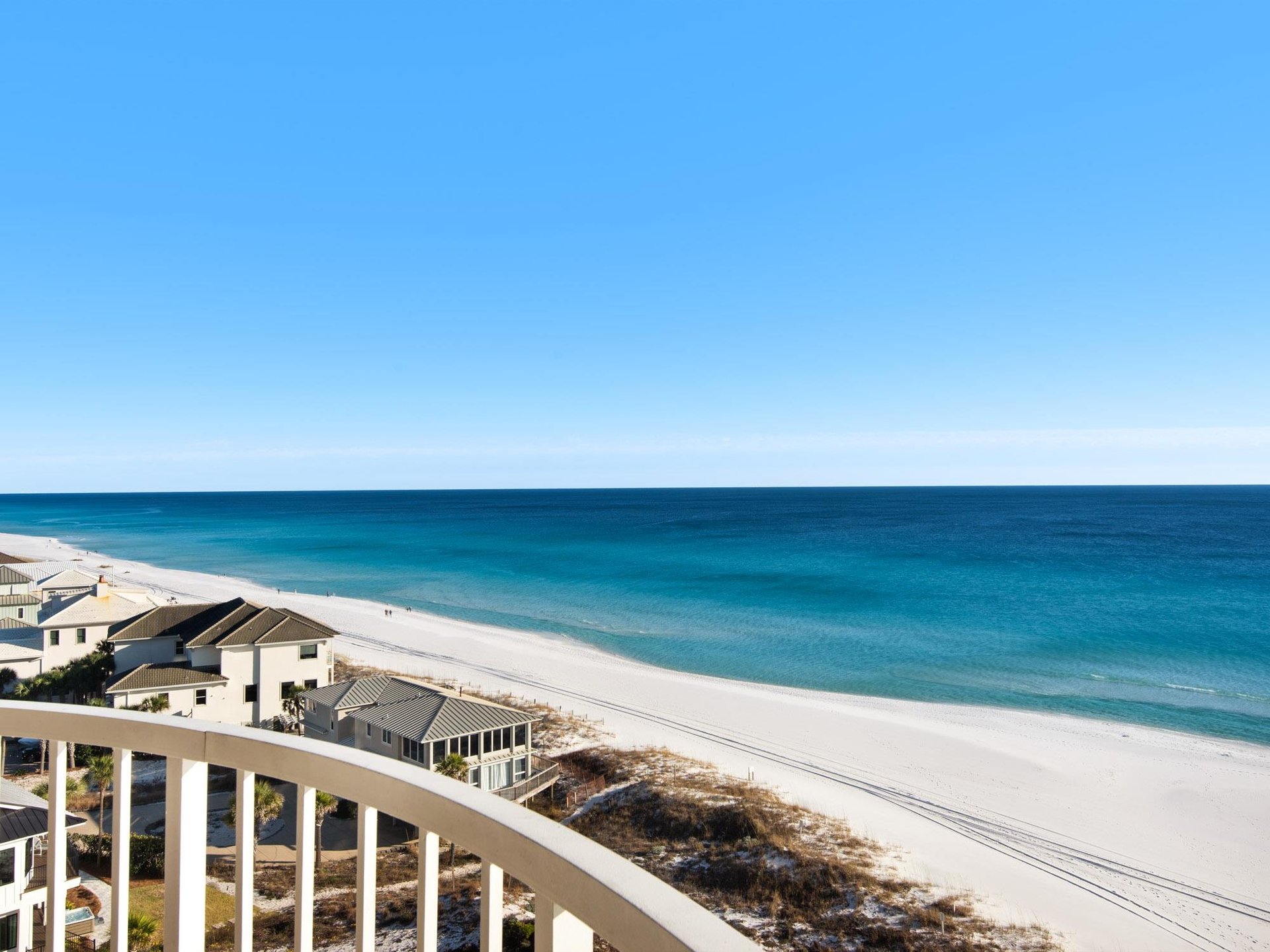 Pristine white sand beach stretches along turquoise waters under clear blue skies, with beachfront homes dotting the coastline.