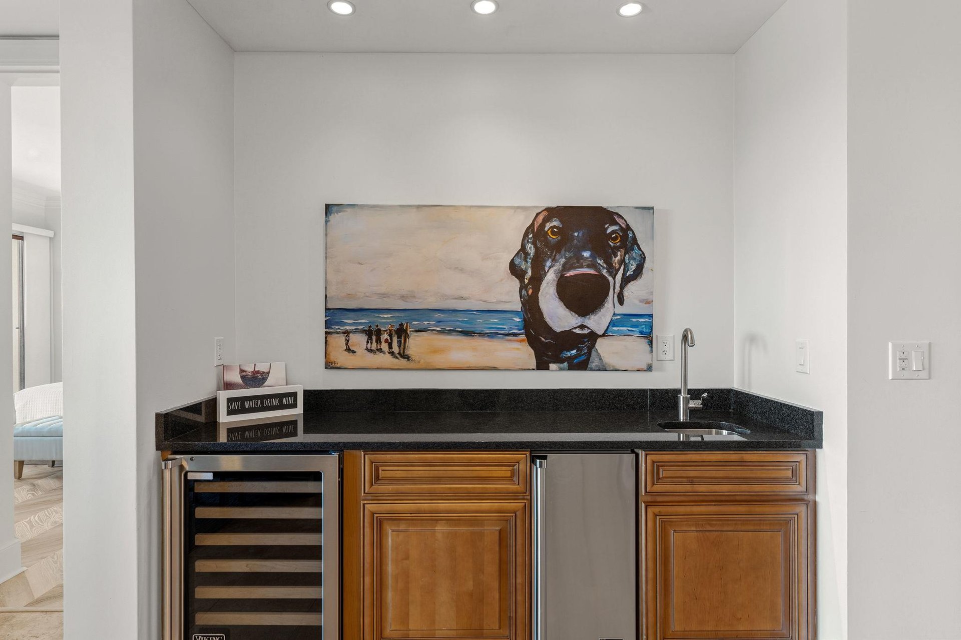 Your stylish wet bar awaits with wine fridge, ice maker, sink, and charming dog artwork—perfect for evening cocktails and coastal conversations.