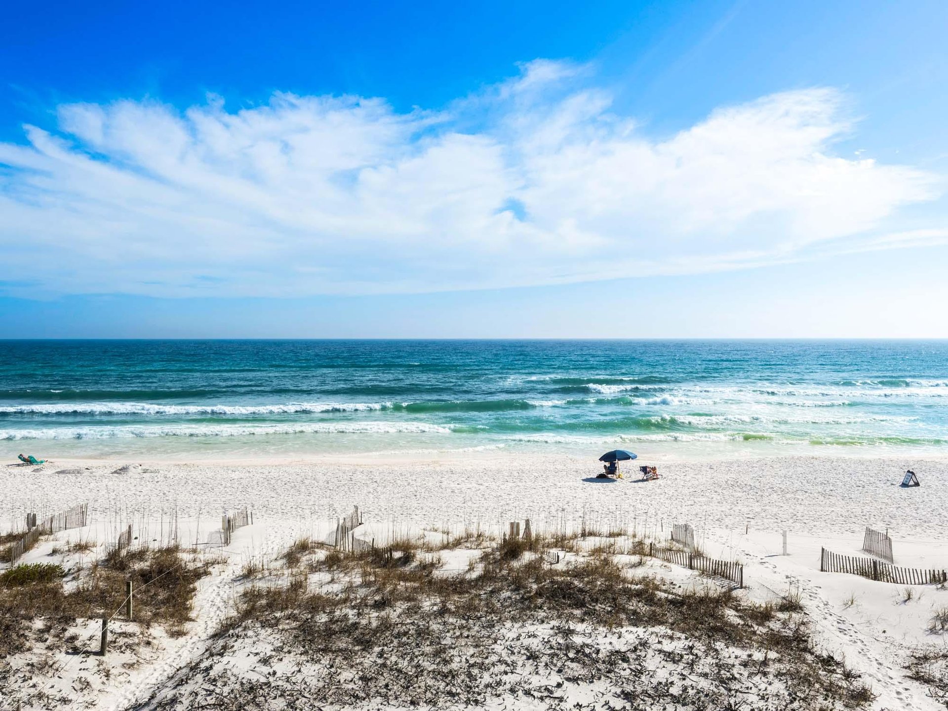 Pristine white sand beach stretches along turquoise waters under dramatic cloudy skies, with gentle waves and scattered beachgoers enjoying the coastal paradise.