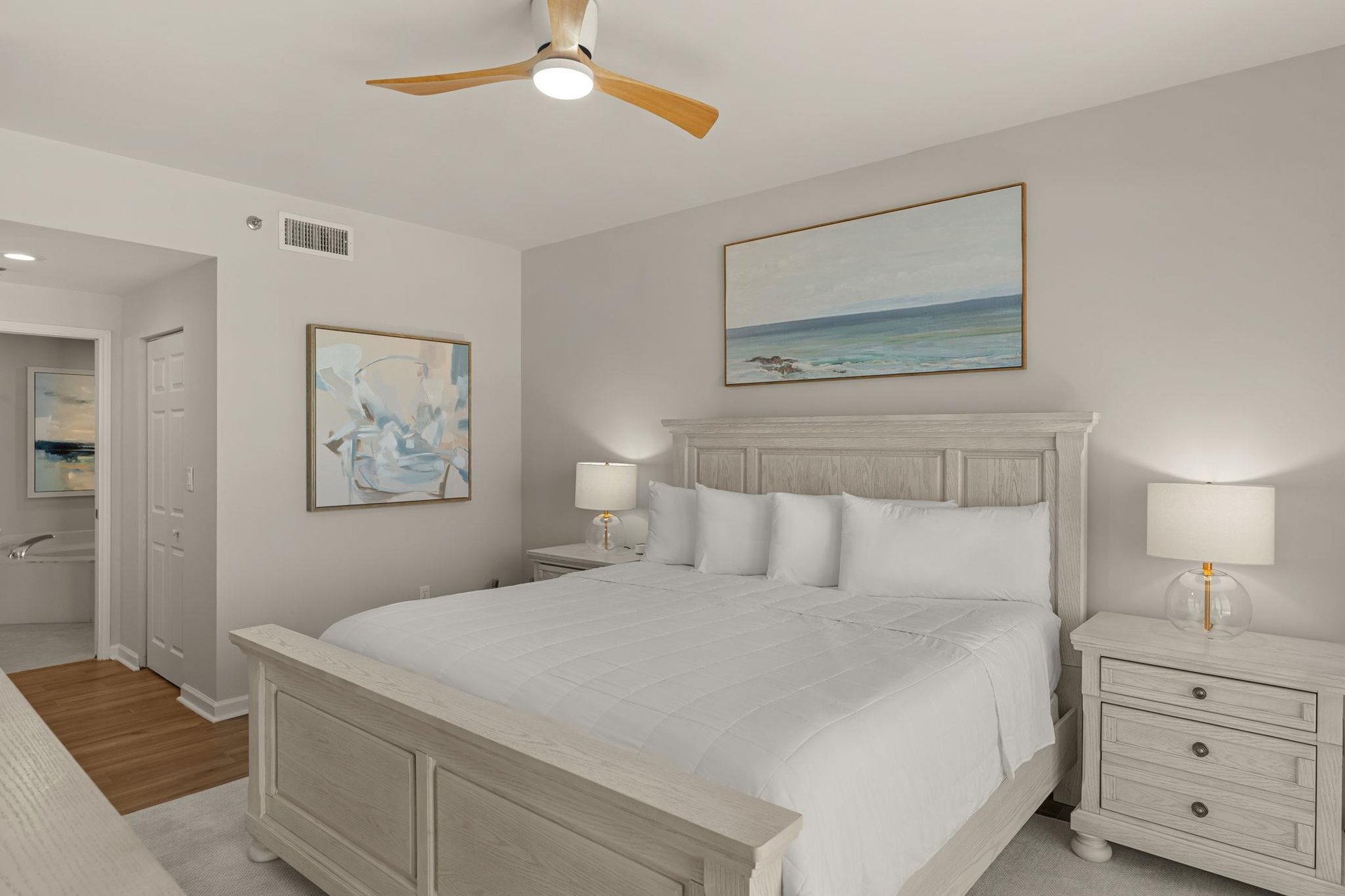 Bedroom 1 with king bed awaits with crisp white linens, coastal artwork, and gentle ceiling fan creating the perfect retreat.