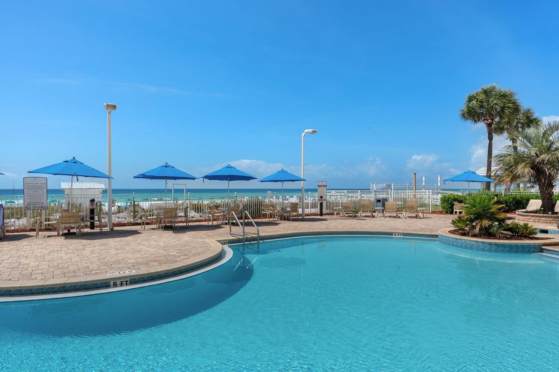 Beachfront pool area with blue umbrellas, lounge chairs, and direct access to pristine sandy shores under clear Florida skies.