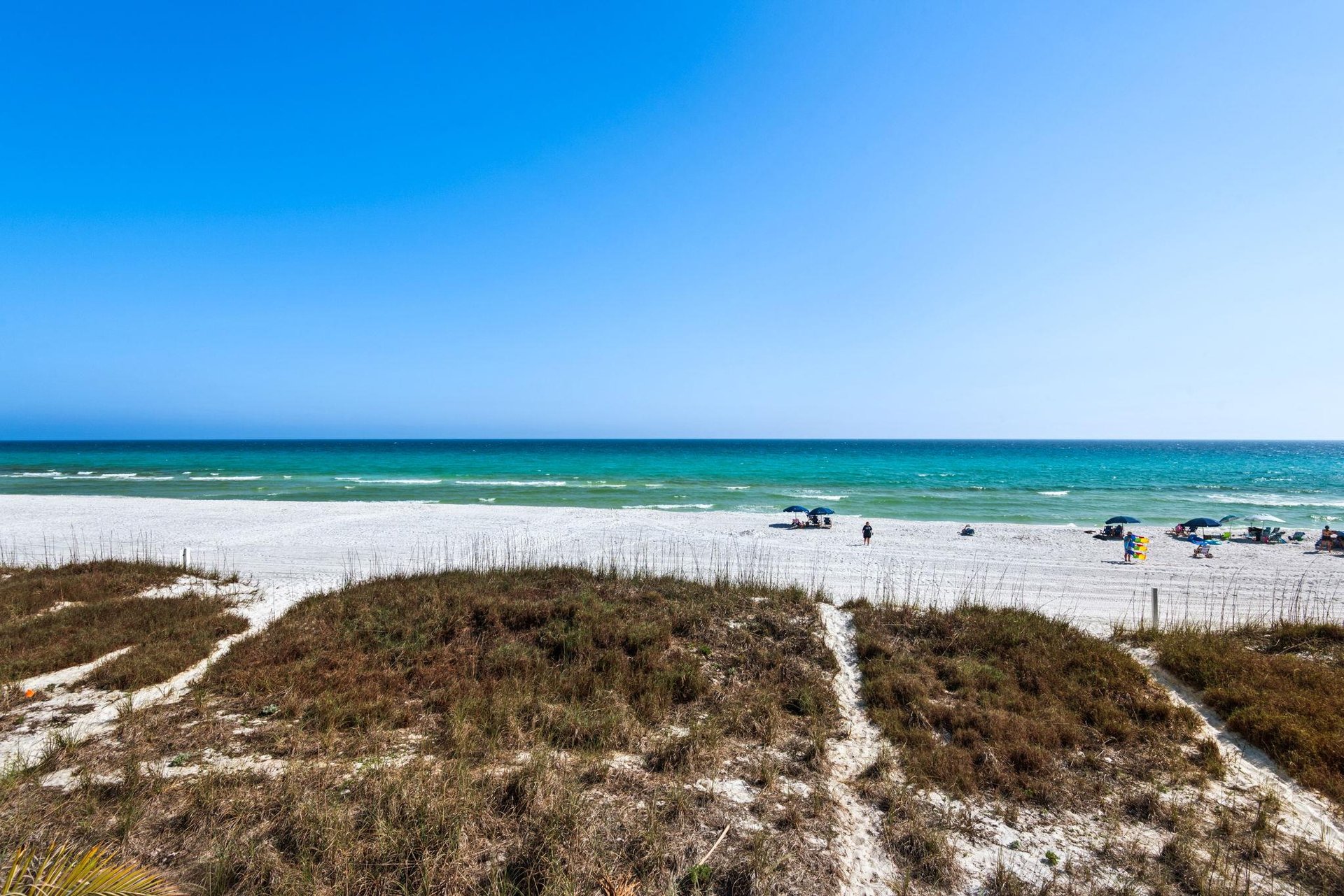 Pristine white sand beach stretches along turquoise waters under clear blue skies, with natural dune vegetation creating coastal pathways.